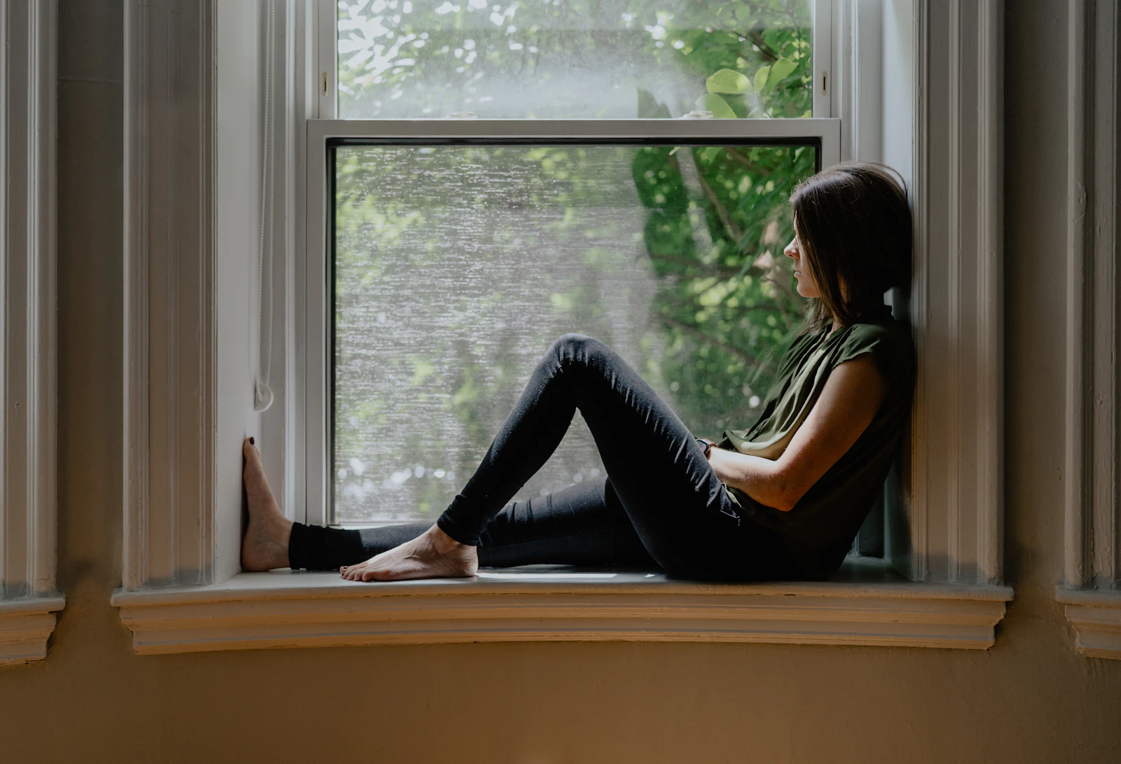 Woman sitting on a window sill in soft natural light, looking out in quiet reflection